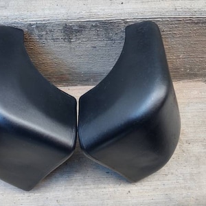 May include: Two black plastic bumper corners for a car. The corners are curved and have a smooth finish.