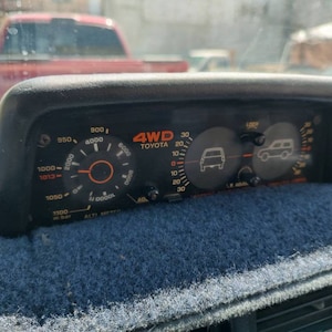 May include: A black dashboard gauge with a 4WD Toyota logo and a dial showing 4WD lock and a dial showing 4WD low.