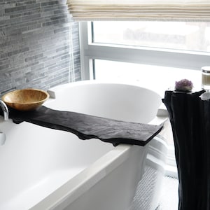 Puede incluir: Una bañera blanca con una bandeja de madera y un cuenco en la parte superior. La bañera está en un baño con una pared de azulejos negros y grises y una ventana. Hay un taburete de madera negro con una vela y un cristal en la parte superior.