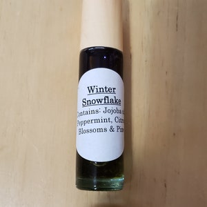 May include: A small glass bottle with a wooden cap and a white label. The label reads "Winter Snowflake" and lists the ingredients: Jojoba Oil, Peppermint, Citrus Blossoms & Pine.