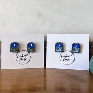 May include: A pair of wooden earrings with a nighttime landscape design. The earrings feature a blue sky with stars and a crescent moon, brown mountains, and a green cactus. The earrings are labeled "Midwest Made".