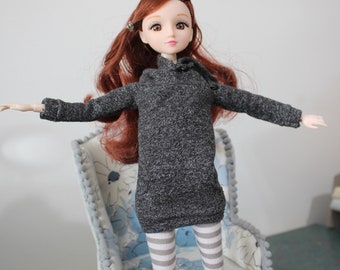 May include: A doll with long brown hair wearing a gray sweater dress and white and gray striped tights. The doll is standing in front of a chair with a floral pattern.