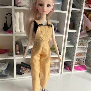 May include: A doll with long blonde hair wearing a yellow and white striped overall dress and black shoes. The doll has brown eyes and pink earrings.