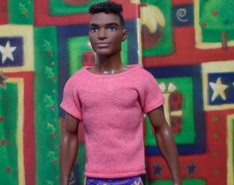 May include: A doll with short, dark brown hair wearing a pink t-shirt and patterned pants. The doll has brown eyes and a light brown complexion. The doll is standing in front of a colorful background.