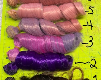 May include: Five bundles of synthetic hair in different colors, including pink, purple, blue, and brown. The hair is arranged in a row from top to bottom, with the numbers 1-6 written next to each bundle.