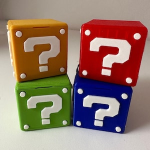Könnte beinhalten: Vier 3D-gedruckte Fragezeichenblöcke in verschiedenen Farben: Gelb, Rot, Grün und Blau. Jeder Block hat ein weißes Fragezeichen auf der Vorderseite.