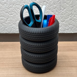 Könnte beinhalten: Ein schwarzer, reifenförmiger Schreibtisch-Organizer mit vier übereinander gestapelten Reifen. Der Organizer ist gefüllt mit Büroartikeln, darunter eine Schere, Stifte und ein weißer Wattestäbchen.