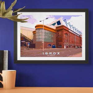 May include: Framed art print of Ibrox Stadium in Glasgow, Scotland. The image features a stylized illustration of the stadium with a red brick facade, orange trim, and blue accents. The text "IBROX GLASGOW" is displayed at the bottom.