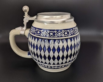 May include: A white ceramic beer stein with a blue and white diamond pattern. The stein has a pewter lid and a handle.