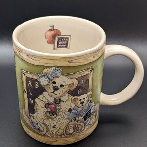 May include: A ceramic coffee mug with a green background and a picture of two teddy bears in a classroom setting. The mug has a brown wooden frame around the image and the text "I can read now" on the inside of the mug.