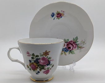 May include: A white teacup and saucer set with a floral design. The cup and saucer have a gold trim around the edges. The floral design features pink, yellow, and blue flowers.