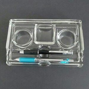May include: A clear glass pen holder with three compartments. The holder has two round compartments and one rectangular compartment. There are two pens in the holder, one black and one blue.