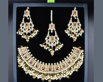 Pode incluir: Um conjunto de joias de noiva de ouro com um colar, brincos e um tikka. O conjunto apresenta pedras grandes de forma quadrada, de cor castanho claro e detalhes de pérolas.