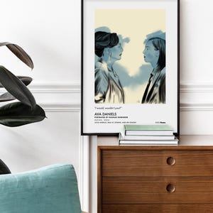 May include: Framed art print featuring a blue-toned illustration of two women facing each other. The artwork includes the text "I would, wouldn't you?" and the name "AVA DANIELS". The print is displayed on a wooden cabinet.