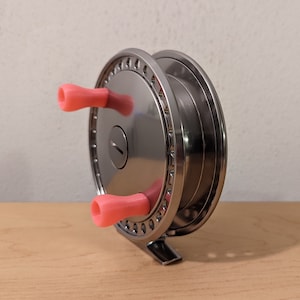 May include: A silver fishing reel with two pink handles. The reel has a silver metal frame and a black spool.