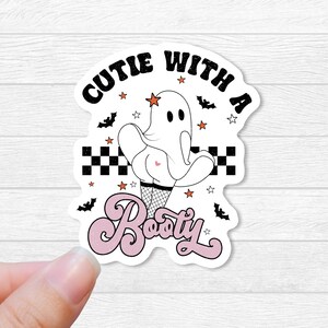 May include: A white ghost with a pink heart on its backside, wearing fishnet stockings and a red star on its head. The ghost is standing on a checkered pattern with bats and stars around it. The text "Cutie with a Booty" is written in pink cursive.