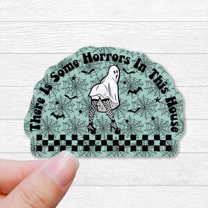 May include: A teal blue sticker with a black and white illustration of a ghost wearing a white dress and fishnet stockings. The ghost is standing on a checkered pattern with a spiderweb background. The text "There is some horrors in this house" is written around the ghost.
