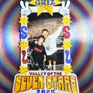 May include: A rectangular frame featuring a photograph of two people on stage, topped with a yellow star labelled GRIZ. The frame sides display blue and pink letters spelling SSL, accented with yellow stars and peace signs. The base reads VALLEY OF THE SEVEN STARS 2026.