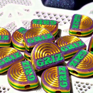 May include: A collection of semi-circular beads featuring a layered design of purple, gold, and green stripes. Each bead is embossed with the text GRIZ in a bold green font, centred over a series of concentric semi-circular gold lines.