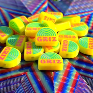 May include: A collection of semi-circular yellow beads featuring a repeating blue rainbow graphic and the text GRIZ printed in pink. Each bead has a small hole for threading, and the surface is smooth with a bright, solid yellow base colour.