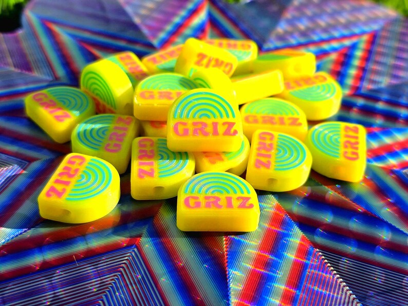 May include: A collection of semi-circular yellow beads featuring a repeating blue rainbow graphic and the text GRIZ printed in pink. Each bead has a small hole for threading, and the surface is smooth with a bright, solid yellow base colour.