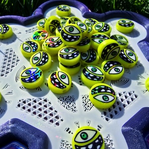 May include: A collection of round, neon yellow beads featuring a black and white graphic of a single eye with a green iris and a wide, toothy grin. The beads are scattered across a white and purple geometric base with a textured, grid-like pattern.