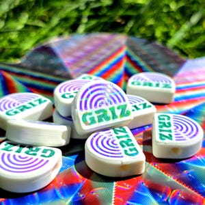 May include: A collection of white, semi-circular beads featuring a purple concentric arc design and the green text GRIZ printed on the flat edge. Each bead has a small hole visible on the side, and they are arranged in a small pile.