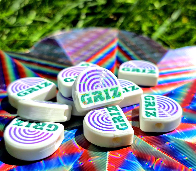 May include: A collection of white, semi-circular beads featuring a purple concentric arc design and the green text GRIZ printed on the flat edge. Each bead has a small hole visible on the side, and they are arranged in a small pile.