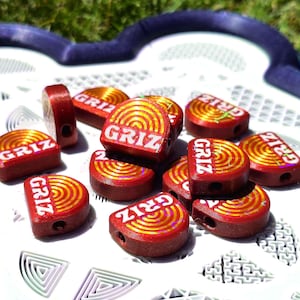 May include: A collection of small, semi-circular red beads featuring a gold-coloured concentric circle pattern and the word GRIZ printed in white. Each bead has a visible hole for threading, and the surface displays a subtle holographic shimmer.