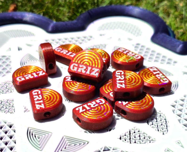 May include: A collection of small, semi-circular red beads featuring a gold-coloured concentric circle pattern and the word GRIZ printed in white. Each bead has a visible hole for threading, and the surface displays a subtle holographic shimmer.