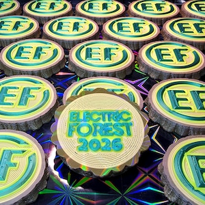 May include: A collection of round, wooden-textured coasters with the letters "EF" in green and yellow. One coaster features the text "ELECTRIC FOREST 2026" in teal and yellow. The coasters are arranged on a reflective, patterned surface.