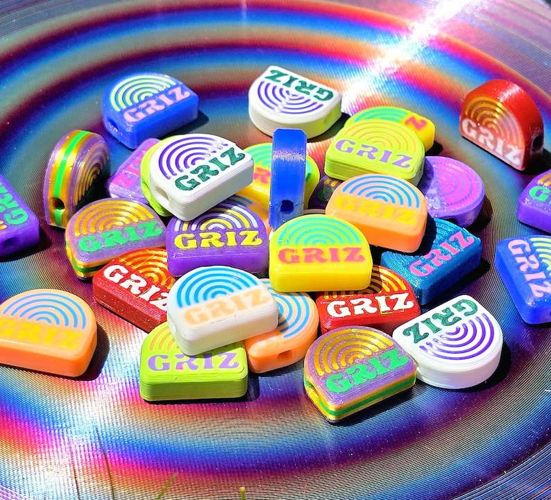 May include: A collection of small, flat, semi-circular beads in various vibrant colours including neon yellow, orange, purple, blue, green, and white. Each bead features a printed rainbow graphic and the text GRIZ in a bold, sans-serif font.
