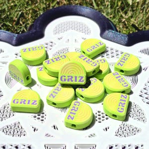 May include: A collection of lime green, semi-circular beads featuring a raised yellow rainbow design and the purple text GRIZ printed across the flat base. Each bead has a small hole on the side, and they are arranged in a scattered pile.