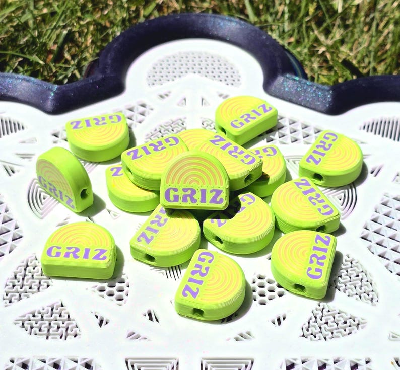 May include: A collection of lime green, semi-circular beads featuring a raised yellow rainbow design and the purple text GRIZ printed across the flat base. Each bead has a small hole on the side, and they are arranged in a scattered pile.