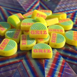 Puede incluir: Un montón de objetos amarillos semicirculares con la palabra "GRIZ" en letras rosas. Cada objeto tiene un diseño de círculos concéntricos turquesa y azules. Los objetos están sobre una superficie colorida con patrones geométricos.