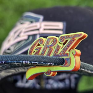 Puede incluir: Un pin colorido impreso en 3D con las letras "GRZ" en una fuente estilizada. El pin presenta un degradado de amarillo, naranja y rojo, y está sujeto a una gorra de béisbol azul oscuro. El fondo está borroso, con hierba verde visible.