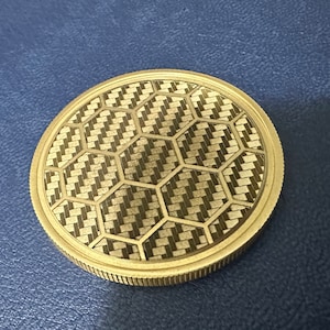 Puede incluir: Una moneda redonda de color dorado con un diseño de panal de abeja.