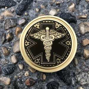 Puede incluir: Una moneda de metal de color dorado con fondo negro y un símbolo médico dorado con alas y una serpiente. La moneda tiene un borde decorativo.