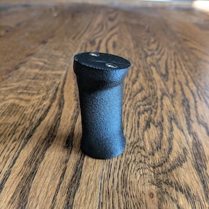 May include: A black, textured, cylindrical object with a slightly tapered waist and a flat top surface. The top features two circular, metallic-colored recessed points. The object has a matte finish and a solid, vertical, upright orientation.