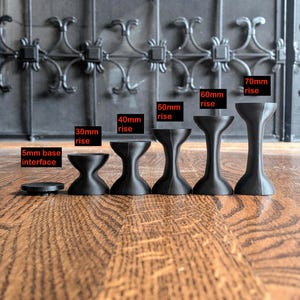 Pu&ograve; includere: Un set di sei supporti neri a forma di clessidra di varie altezze. Le etichette indicano una base di 5mm, seguite da opzioni di 30mm, 40mm, 50mm, 60mm e 70mm. Ogni pezzo presenta una sezione centrale concava e una superficie superiore piatta.