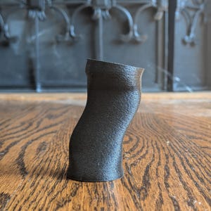 Pu&ograve; includere: Un piccolo oggetto cilindrico nero stampato in 3D con un corpo leggermente curvo e asimmetrico e una superficie ruvida. L'articolo presenta un'apertura superiore cava e una base piatta, rimanendo in posizione verticale con una forma organica.