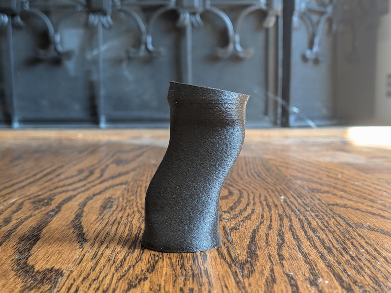 Pu&ograve; includere: Un piccolo oggetto cilindrico nero stampato in 3D con un corpo leggermente curvo e asimmetrico e una superficie ruvida. L'articolo presenta un'apertura superiore cava e una base piatta, rimanendo in posizione verticale con una forma organica.