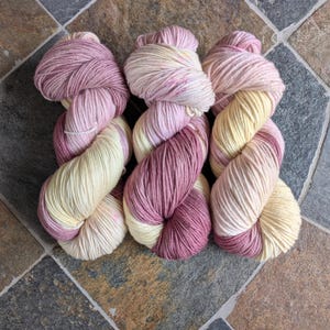 May include: Three skeins of hand-dyed yarn in a color palette of cream, light pink, and deep rose. The yarn is twisted into a skein shape, showing the color variations. The yarn is likely for knitting or crochet projects.