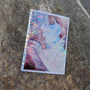 May include: A holographic sticker featuring a detailed illustration of a blue and white creature with a crown-like headpiece. The creature has a long, slender body and is depicted in a pose that suggests movement. The background is a watercolor wash of red and purple.