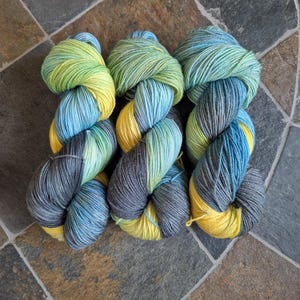 May include: Three skeins of colorful yarn in shades of blue, green, yellow, and gray. The yarn is arranged in a twisted pattern, showcasing the color variations. The skeins are on a stone tile surface.