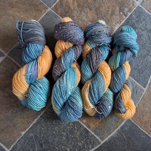 May include: Four skeins of hand-dyed yarn in a variety of colors. The yarn is a mix of blue, teal, gold, and gray. The yarn is arranged in a twisted pattern.