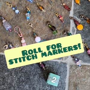 Puede incluir: Una colección de marcadores de punto de colores con cuentas y abalorios. Un dado verde con el número 2 está en primer plano. El texto "Roll for Stitch Markers!" está en una pancarta amarilla.