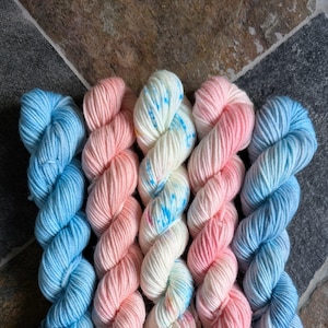 May include: Five twisted skeins of yarn arranged side by side. The outer skeins are light blue, the second and fourth are soft peach, and the center skein is cream-colored with bright blue and pink speckles throughout the twisted fiber strands.