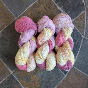 May include: Three skeins of hand-dyed yarn in shades of pink, lavender, and yellow. The yarn is arranged in a twisted pattern, showcasing the colour variations. The skeins are on a tiled surface, providing a neutral backdrop for the vibrant colours.