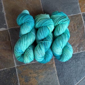 Peut inclure: Trois écheveaux de fil teints à la main dans des tons de sarcelle et de turquoise. Le fil est disposé en rangée, chaque écheveau présentant un dégradé de couleurs. Le fil est doux et duveteux, parfait pour les projets de tricot ou de crochet.